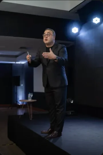 Marcos Dantas em pé no palco, falando para uma plateia.
